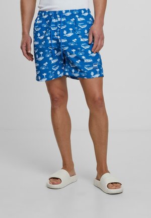 Shorts de bain bleu avec un motif tropical blanc, taille élastique avec cordon de serrage, et longueur au-dessus du genou. Portés avec des sandales blanches.