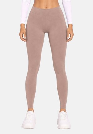 Femme portant un legging taupe taille haute, un haut blanc à manches longues et des baskets blanches, debout devant un fond clair uni.