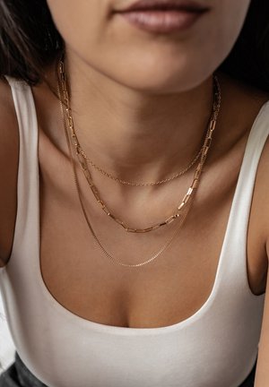 Drie gelaagde gouden kettingen: één met langwerpige rechthoekige schakels, één met kleine ronde schakels en een dunne ketting. Draag over een witte tanktop.