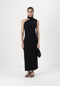 Haut noir à une épaule avec une texture froncée, associé à une jupe longue et ajustée noire. Le modèle tient un petit sac à main noir et porte des sandales à lanières.