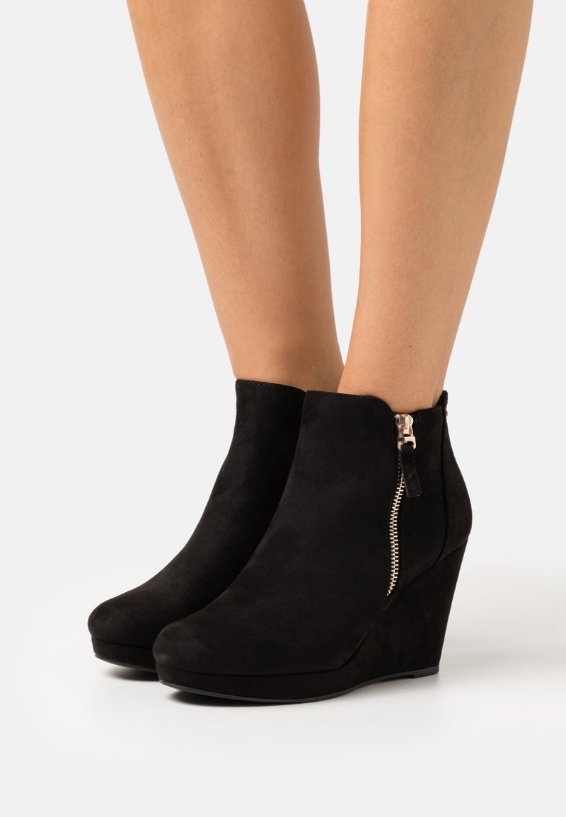 Jambes portant des bottines compensées en daim noir avec des fermetures éclair latérales sur un fond blanc.