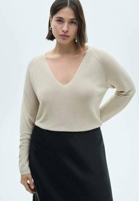 Beige geribbeld sweater met een diepe V-hals en lange mouwen, gecombineerd met een zwarte rok. Zachte textuur en getailleerde silhoutten.