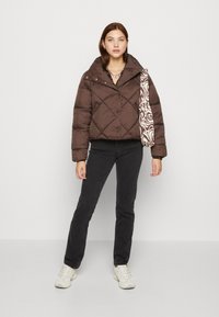 Doudoune brune avec un col montant et un design matelassé, associée à un jean noir et un sac à motifs. Le mannequin porte des baskets.