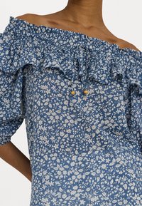 Modrá květinová top s odhalenými rameny a rýhovaným výstřihem; má stahovací šňůrky s žlutými korálky; látka má texturovaný, lehký vzhled.