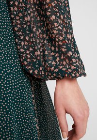 Dunkelgrünes Kleid mit kontrastierenden Punkten und floralen Mustern in Pink und Weiß. Der Stoff ist durchsichtig mit einer strukturierten Oberfläche.