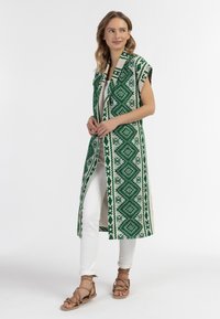 Zaļš rakstains kimono stila veste ar dimanta motīviem, atvērta priekšpuse, īsām piedurknēm, valkāta virs baltas blūzes un pieguļošām baltām biksēm.