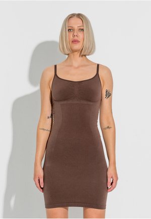 Femme blonde avec une coupe au carré porte une robe marron ajustée sans manches, debout sur fond blanc avec des tatouages visibles sur les bras.