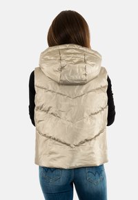 Gilet beige bouffant avec capuche, doté de coutures en chevron et d'une finition brillante. Porté sur un haut noir, vu de dos.