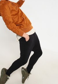 Sudadera de color óxido con un bolsillo frontal, superpuesta a una camisa blanca, combinada con pantalones ajustados negros y zapatillas deportivas verdes.