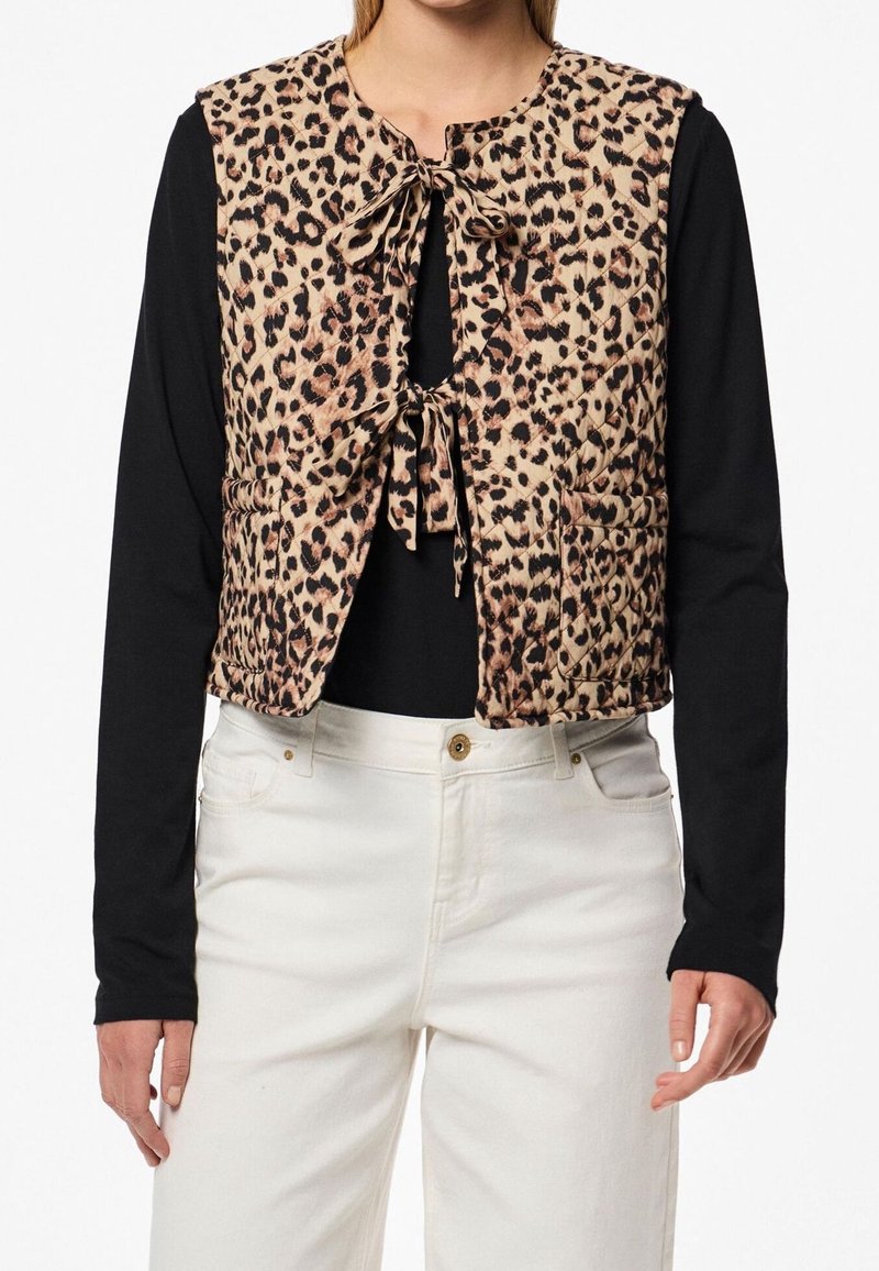 Femme portant un gilet sans manches beige et noir à imprimé léopard, noué devant, par-dessus une chemise noire à manches longues et un pantalon blanc.