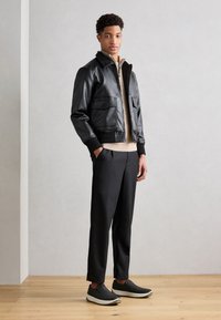 Veste bomber en cuir noir avec deux poches avant, un pull zippé beige en dessous, un pantalon noir et des chaussures gris à enfiler.