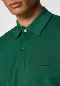 Grønn polo skjorte laget av mykt stoff med knappete krage, prydet med brodert "STUDENTER" i svart på forsiden.
