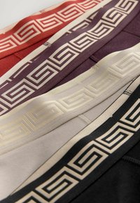 Vier Paare Taillenbandstoffe in verschiedenen Farben—rot, dunkelrot, beige und schwarz—mit einem geometrischen Muster in Gold entlang der Oberkante.