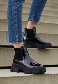 Bottines en cuir verni noir avec une semelle épaisse, des panneaux latéraux élastiques et des détails texturés. Portées avec un jean en denim effiloché sur des escaliers.