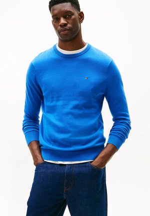 Homme portant un pull bleu vif avec un petit logo sur la poitrine et un jean bleu foncé, les mains dans les poches, sur un fond blanc.