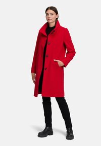 Vrouw staat in een rode button-up jas met hoge kraag, zwarte broek en zwarte stevige laarzen, met één hand in de jaszak tegen een witte achtergrond.