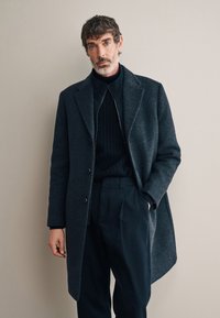 Cappotto di lana grigio scuro con semplici rever, chiusura con un solo bottone e taglio dritto, indossato sopra un maglione a costine scuro e pantaloni.