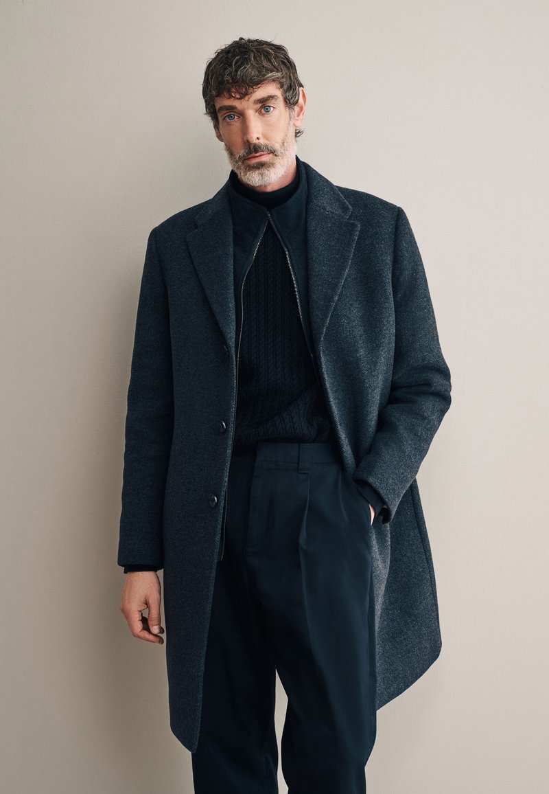 Cappotto di lana grigio scuro con semplici rever, chiusura con un solo bottone e taglio dritto, indossato sopra un maglione a costine scuro e pantaloni.