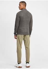 Grå ribbad tröja med hög krage, tillsammans med ljus khaki-byxor. Modellen bär vita sneakers med bruna detaljer. Båda plaggen har en figurnära design.