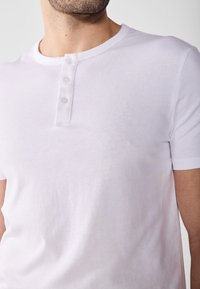 Bílý bavlněný henley tričko s krátkým rukávem, s kulatým výstřihem a třemi knoflíky na přední straně. Hladká, lehká textura.