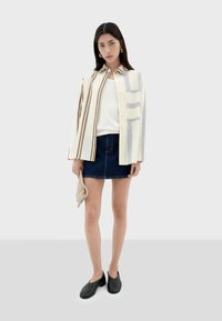Gestreifte Button-Down-Bluse in Creme, Braun und Blau. Weißes Oberteil, dunkler Jeansrock, schwarze Schuhe. Hält eine beigetönige Stricktasche.