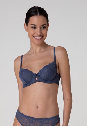 Donna sorridente che indossa reggiseno e mutandine in pizzo blu navy abbinate su uno sfondo semplicemente grigio chiaro.