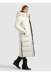 Ilgas baltas pūkinis paltas su siūtu raštu, turi gobtuvą, juodus užtrauktukus ir šonines kišenes. Derinamas su juodais, iki čiurnos aukščio, wedge tipo batais.