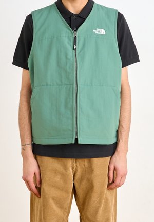 Veste sans manches verte zippée avec le logo The North Face portée sur un polo noir et un pantalon en velours côtelé marron.