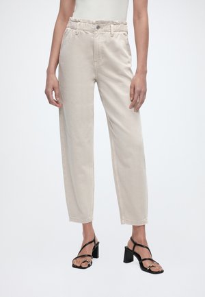 Osoba ubrana w beżowe spodnie o wysokim stanie i luźnym kroju, w zestawieniu z czarnymi sandałami na słupku z paskami, stojąca na tle jednolitego, jasnego tła.