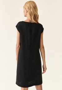 Blonde Frau mit schulterlangem Haar, die ein schwarzes, kurzärmliges, knielanges Kleid trägt und mit dem Rücken zur Kamera steht, wobei ein Tattoo auf ihrem oberen Rücken zu sehen ist.