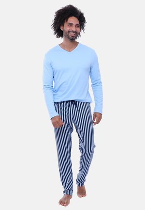 Uśmiechnięty mężczyzna o kręconych włosach, ubrany w jasnoniebieską koszulę z długim rękawem oraz granatowo-białe pionowo paskowane spodnie, idący boso.