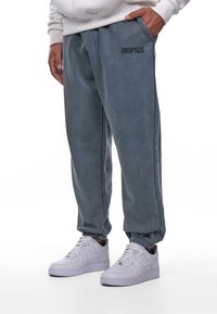 Grå sweatpants lavet af blødt stof, med elastisk talje, sidelommer og et sort "DROPSIZE" logo på forsiden.