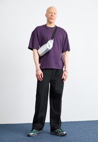 Lila kortärmad t-shirt, svarta byxor, silverfärgad axelväska och gröna och grå sneakers. Neutral bakgrund och matt golv.