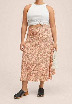 Žena v bílém uzlovaném tílku bez rukávů, oranžové květované midi sukni, černých dreváčcích a držící bílou pletenou kabelku.