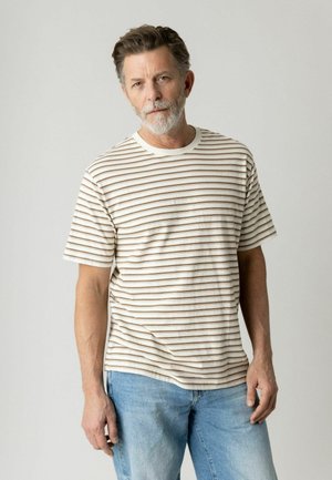 Hombre de mediana edad con cabello y barba gris, vestido con una camiseta de rayas beige y blanca y pantalones vaqueros azul claro, sobre un fondo liso.
