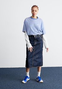 T-shirt a righe blu e bianche a maniche corte indossato sopra una camicia bianca a maniche lunghe, abbinato a una gonna a matita in denim scuro e sneaker blu.