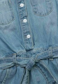 Ljusblå denimplagg med vita knappar fram, bröstfickor och ett knytet tygbälte i midjan.