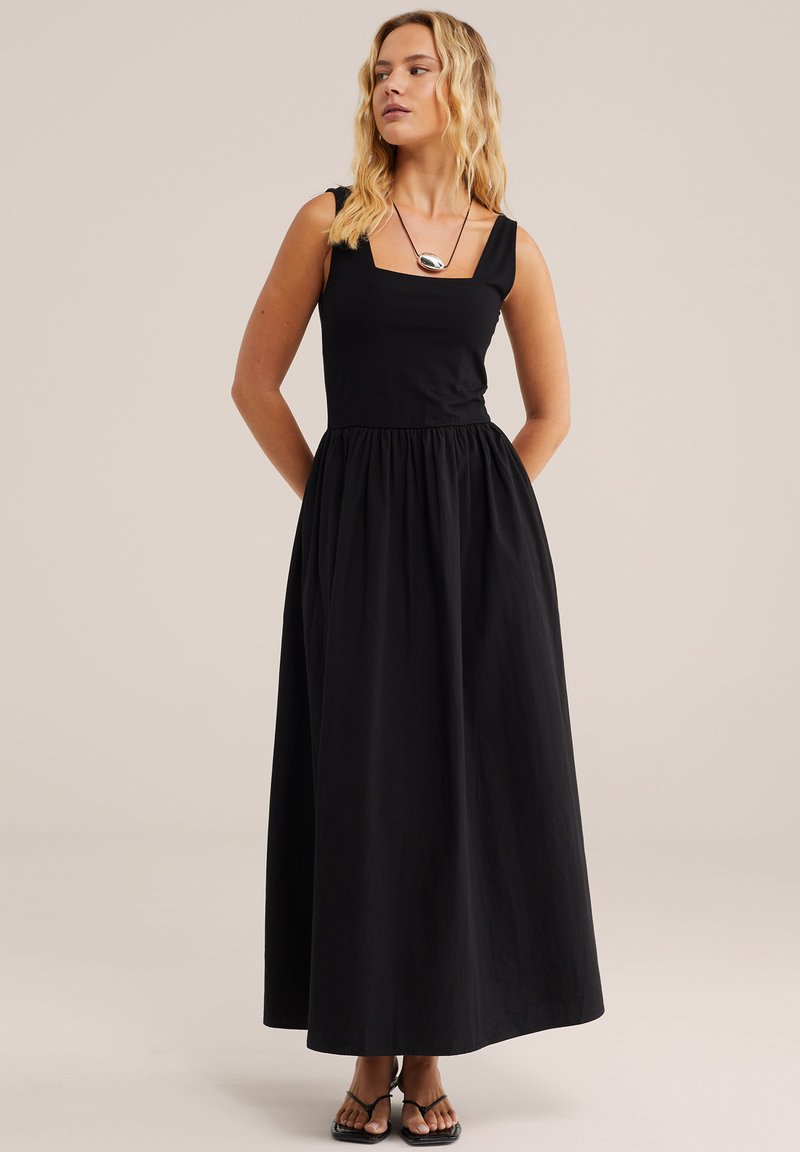 Mujer de cabello rubio con un vestido maxi negro sin mangas de escote cuadrado y sandalias negras, de pie con las manos detrás de la espalda.