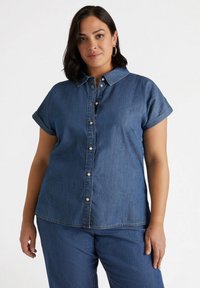 Camisa de mezclilla en azul oscuro, con cuello tipo solapa, mangas cortas enrolladas, cierre frontal con botones y sutilezas en las costuras.