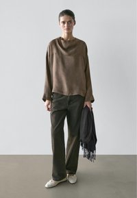 Vrouw staand tegen een effen muur, gekleed in een loszittende bruine lange mouwen top, donker wijde broekspijpen, witte schoenen, met een zwarte sjaal met franjes in haar rechterhand.