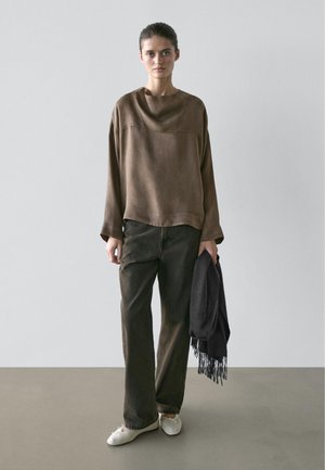 Woman standing against plain wall wearing loose brown long-sleeve top, dark wide-leg pants, white shoes, holding black fringed scarf in right hand.