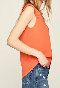 Ljusa orange ärmlös blus med ett rynkat halslinning, gjord av lätt tyg, i kombination med slitna blå denimshorts.