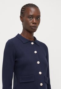 Tumedast sinine kudumiga kardigan krae ja kuue hõbedase nööbiga, millel on kaks eesmise taskut ja räbaldunud tekstuur.