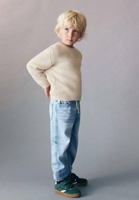 Beige geripptes Sweatshirt, hellblaue weite Jeans mit Kordelzug und grüne Turnschuhe mit einer braunen Sohle, getragen von einem Kind mit kurzen blonden Haaren.