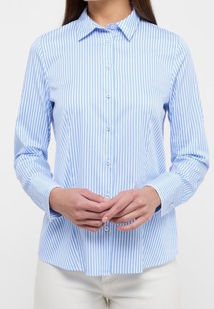 Woman wearing a long-sleeve, light blue and white vertically striped button-up shirt, hands clasped in front, white pants visible.