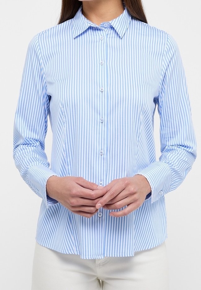 Femme portant une chemise à manches longues rayée verticalement bleu clair et blanc, mains jointes devant, pantalon blanc visible.