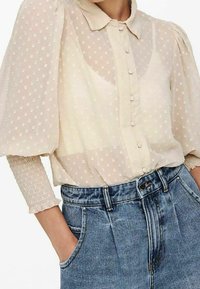 Przezroczysta beżowa bluzka w kropki, z bufiastymi krótkimi rękawami i zapięciem na guziki z przodu, połączona z wyblakłymi niebieskimi szortami dżinsowymi z wysokim stanem.