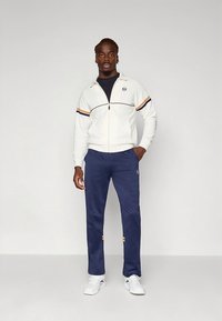 Hvit jakke med glidelås med marineblå og oransje striper, kombinert med marineblå bukser med logo-accenter og hvite joggesko. Enkel, sporty design.