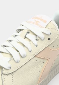 Cream leather sneaker with white laces and a peach accent logo. Features a textured surface and a rubber sole with gray detailing.