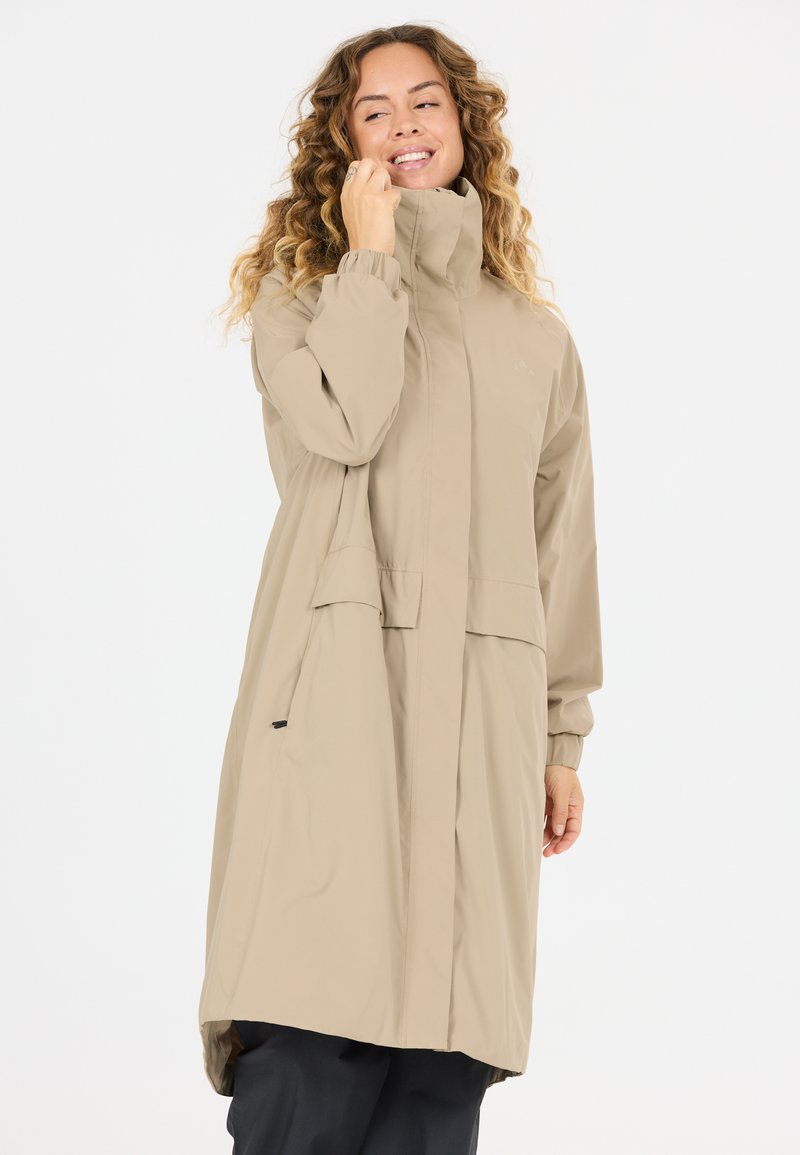 Nainen, jolla on kiharat hiukset ja joka käyttää beigeä korkeakauluksista pitkää takkia, hymyilee ja koskettaa kaulusta yksinkertaisen vaalean taustan edessä.